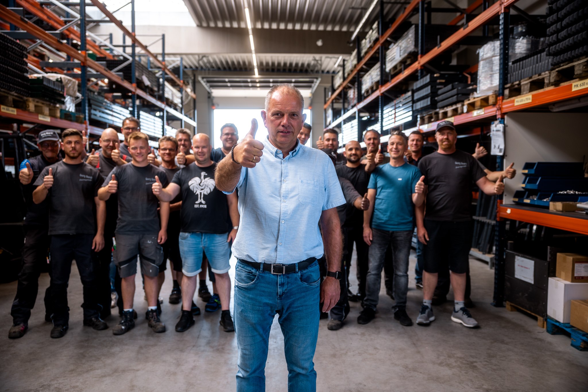 Gruppe von Menschen in Lagerhalle, vorne ein Mann mit blauem Hemd und Daumen hoch, dahinter mehrere Personen in Freizeitkleidung ebenfalls mit Daumen hoch