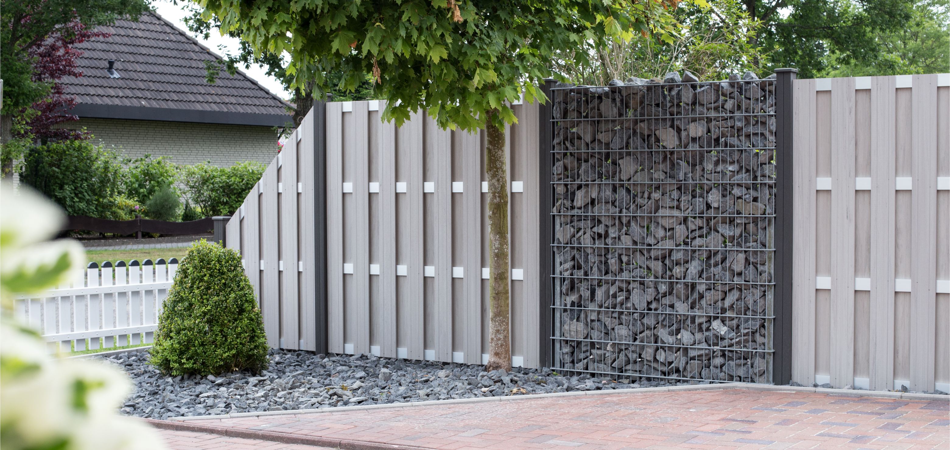 Moderner Zaun mit senkrechten hellen Paneelen und einem Gabionen-Sichtschutz mit grauen Steinen in einem Garten mit Baum und Hecke