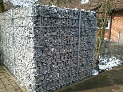 System Gabionensteine - Großer rechteckiger Gabionenkorb gefüllt mit grauen Steinen steht auf gepflastertem Boden vor einem Haus mit Satteldach