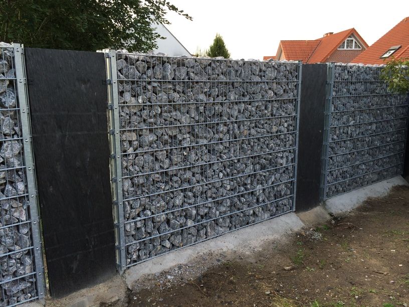 System Gabionensteine - Gabionenwand mit grauen Steinen in Metallrahmen zwischen schwarzen Sichtschutzwänden vor Wohnhäusern