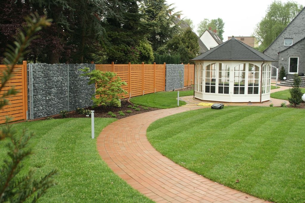 System Gabionensteine -Garten mit geschwungenem roten Ziegelweg, grünem Rasen, Holzzaun und einem weißen Pavillon mit dunklem Dach