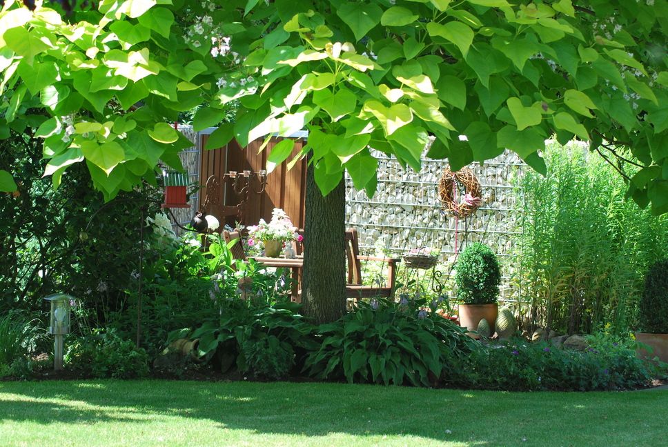 System Gabionensteine - Garten mit großem Baum, Blumenbeeten, Gartenmöbeln und einer Steinmauer im Hintergrund