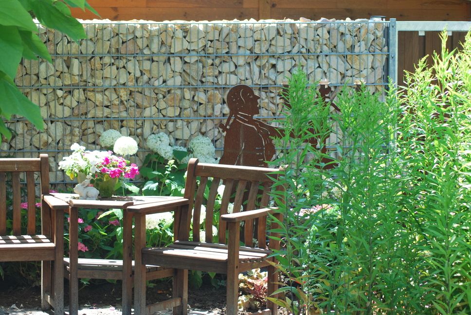 System Gabionensteine - Garten mit Holzstühlen, Tischen, Topfpflanzen und einer Metallskulptur vor einer Gabionenwand