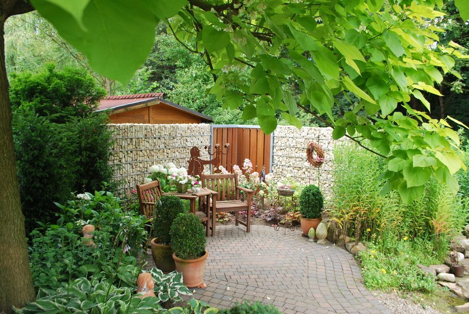 System Gabionensteine - Gepflasterte Gartenecke mit Holzbank, Tisch, Blumentöpfen und grünem Laub im Vordergrund