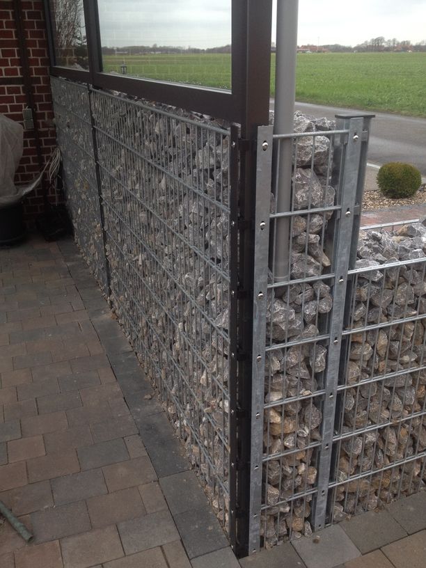 Gabionenwand aus Metallgittern, die mit Steinen gefüllt sind, neben einem gepflasterten Weg und einem Haus mit rotem Backsteinmauerwerk