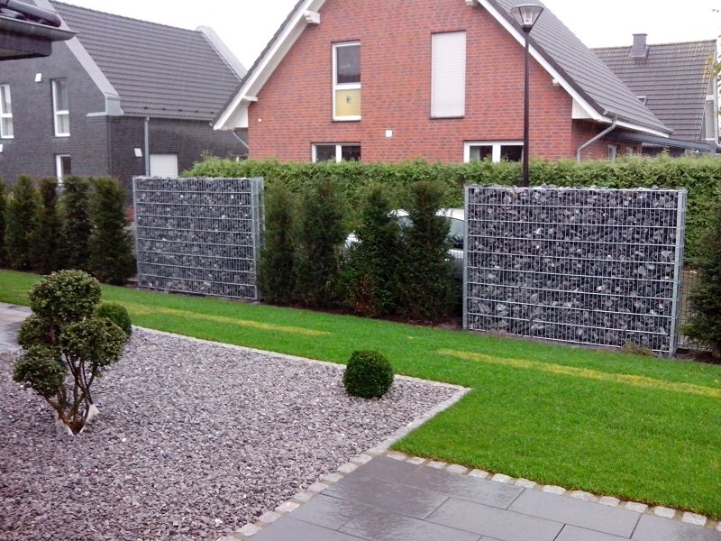 System Gabionensteine - Garten mit Rasen, Kiesfläche, zwei gabionenartigen Steinmauern und Hecke vor einem roten Backsteinhaus mit grauem Dach
