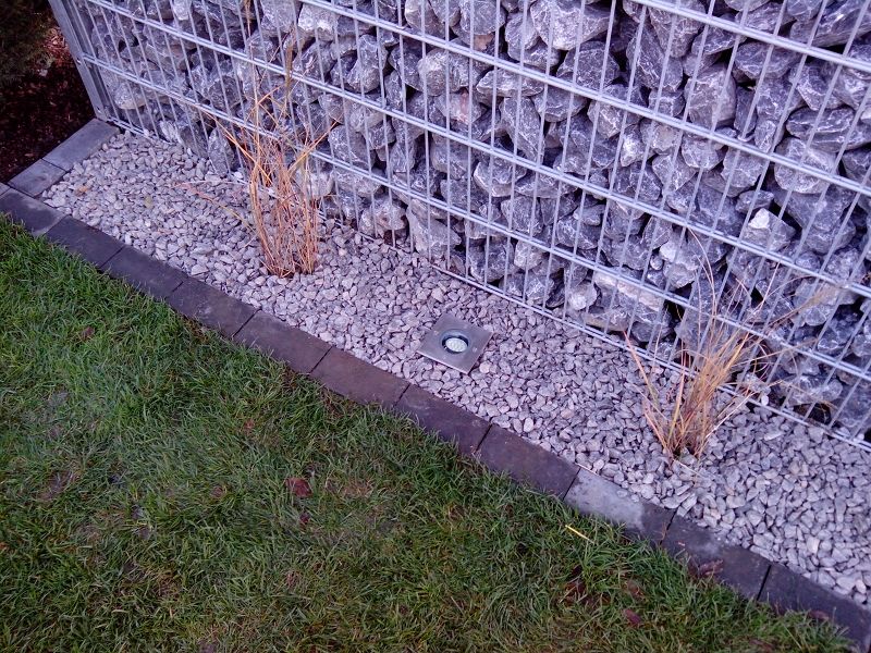 System Gabionensteine - Gabionenwand mit grauem Kiesbett davor, in dem zwei vertrocknete Pflanzen und eine Bodeneinbauleuchte stehen, angrenzend an einen Rasenstreifen