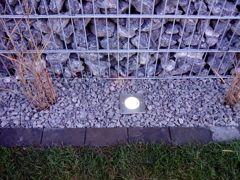 System Gabionensteine - Beleuchteter Bodeneinbaustrahler in einem Kiesbett vor einer Gabionenwand aus Steinen und einem kleinen Grasstreifen
