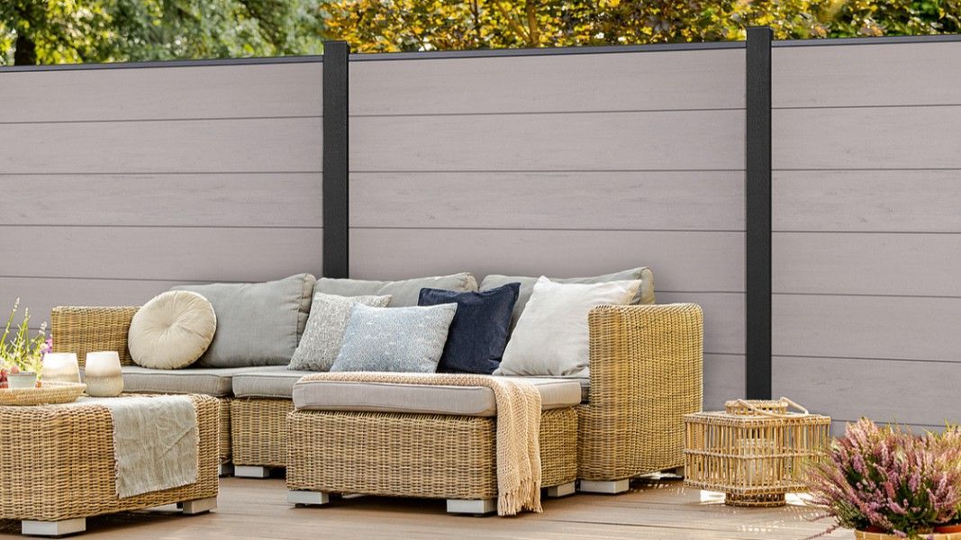 Ecksofa aus geflochtenem Rattan mit hellen Polstern und mehreren Kissen vor einer grauen Sichtschutzwand