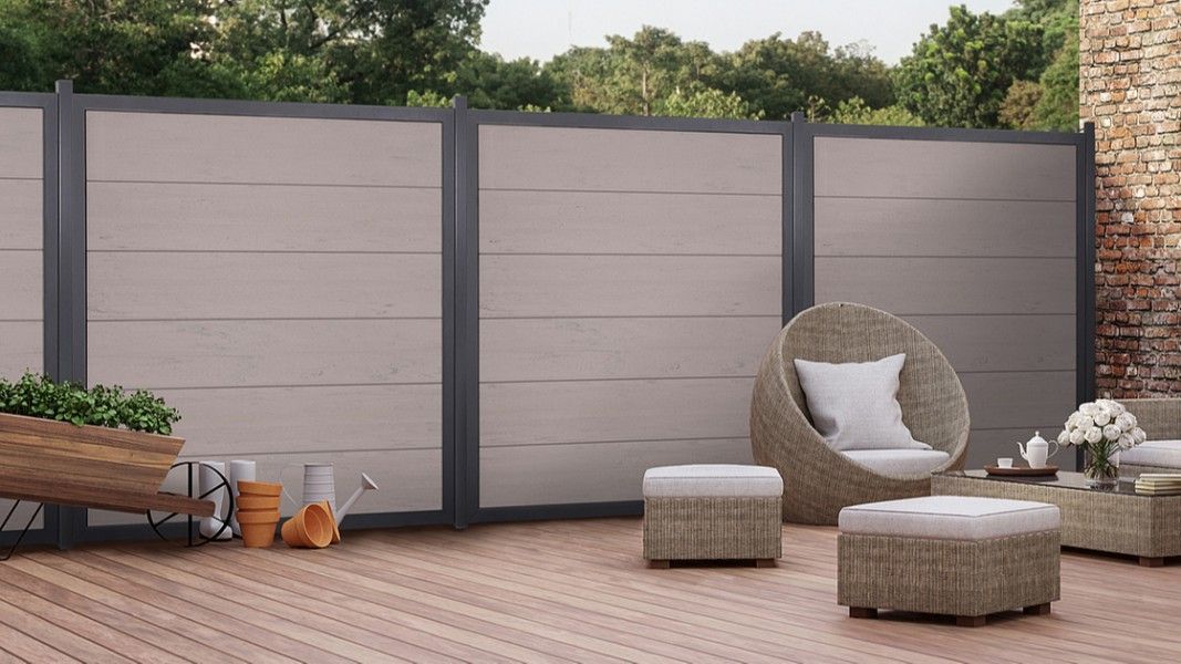 Groja Viento - Terrasse mit Holzlattenboden, grauer Sichtschutzwand, runder Korbsessel mit weißem Kissen und zwei quadratischen Korbhockern