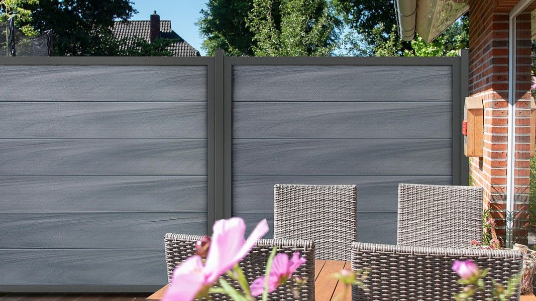 Groja Viento - Garten mit grauer Sichtschutzwand, zwei geflochtenen Stühlen und verschwommenen rosa Blumen im Vordergrund