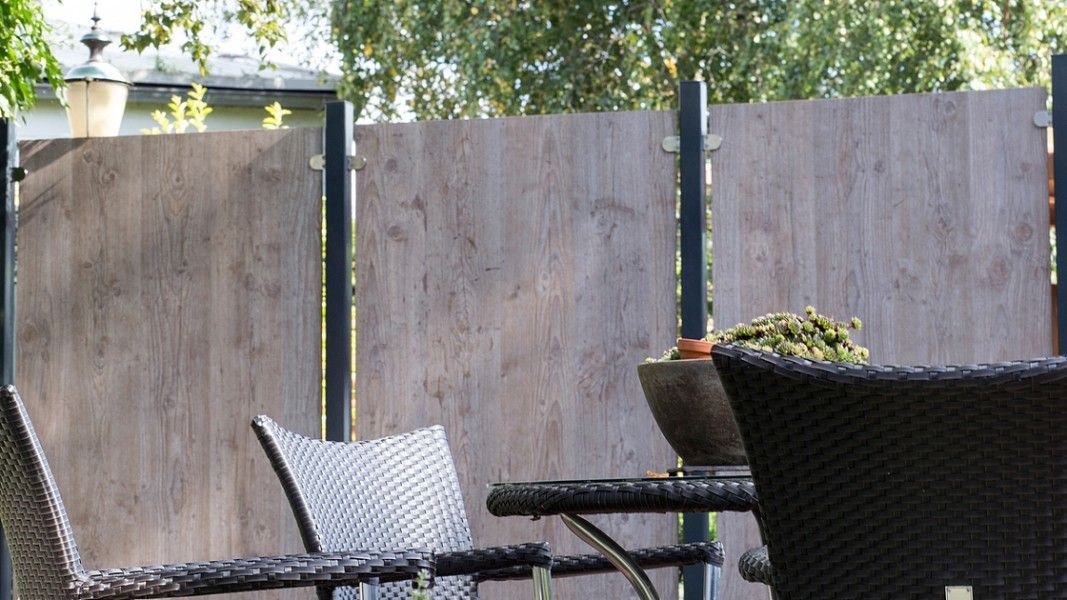 Groja Premo - Gartenbereich mit geflochtenem Stuhl und Tisch vor einer Holzsichtschutzwand und Bäumen im Hintergrund